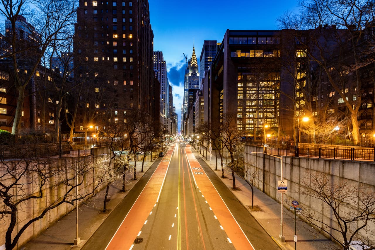 A city street in Murray Hill, Manhattan is flanked by tall buildings and illuminated at dusk. The road is mostly empty, with streetlights casting a warm glow. The Chrysler Building is visible in the distant background.