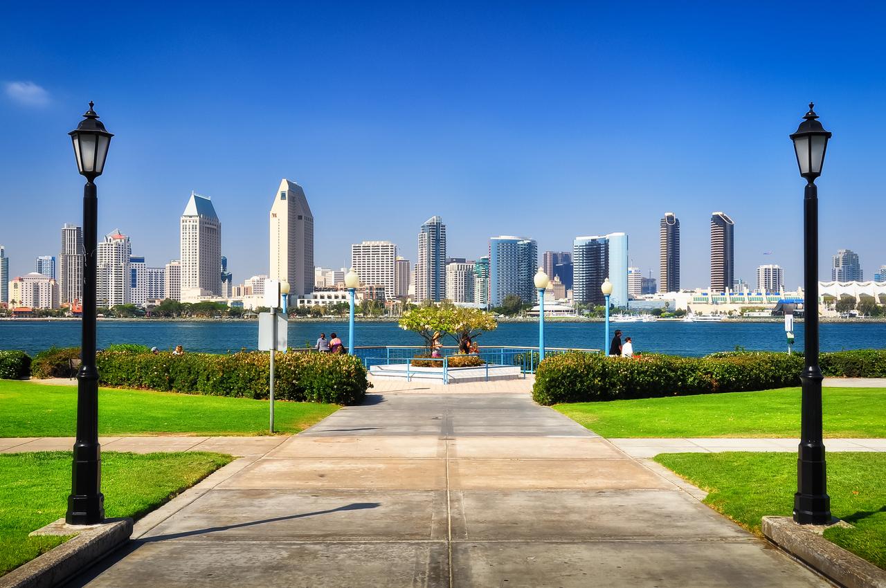 A view of a city skyline featuring modern high-rise buildings across a body of water, framed by two street lamps and green lawns on either side of a paved pathway.