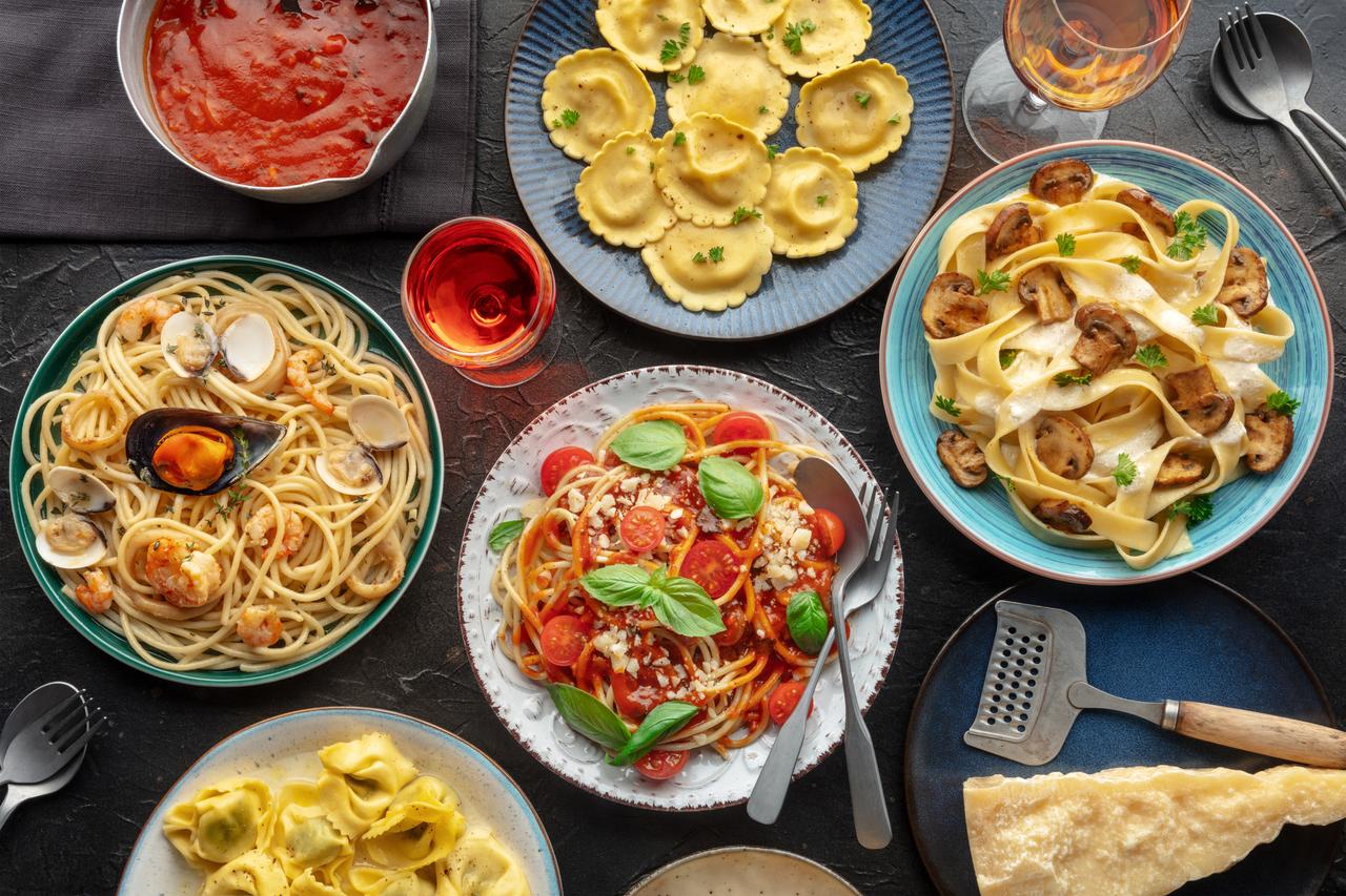 An assortment of pasta dishes, including ravioli, spaghetti with seafood, fettuccine with mushrooms, and tortellini, served with sauces and garnished with basil. Multiple utensils and glasses of drinks nearby. Experience this culinary delight on our exclusive food tour.