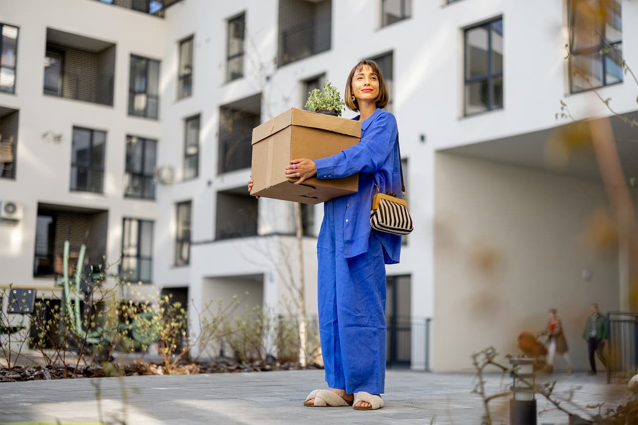 A woman in a blue jumpsuit holding a box and purse, standing in an apartment complex courtyard, facing the camera.