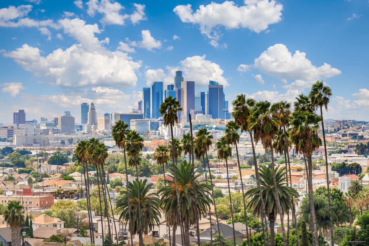 Downtown Los Angeles skyline with tall buildings and palm trees in the foreground, under a blue sky with scattered clouds.